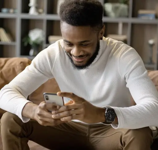 A man sitting on a couch, smiling and looking at his smartphone screen