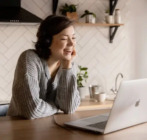 A woman wearing a grey sweater, is smiling and engaging in a video call on her laptop in a modern kitchen