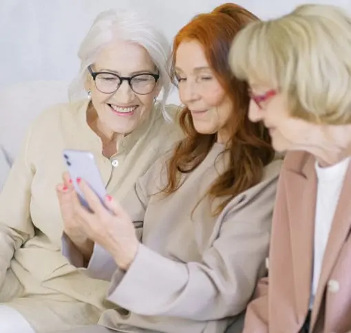 Three smartly-dressed women in their 60s gathered around one smart phone and smiling at what they see.